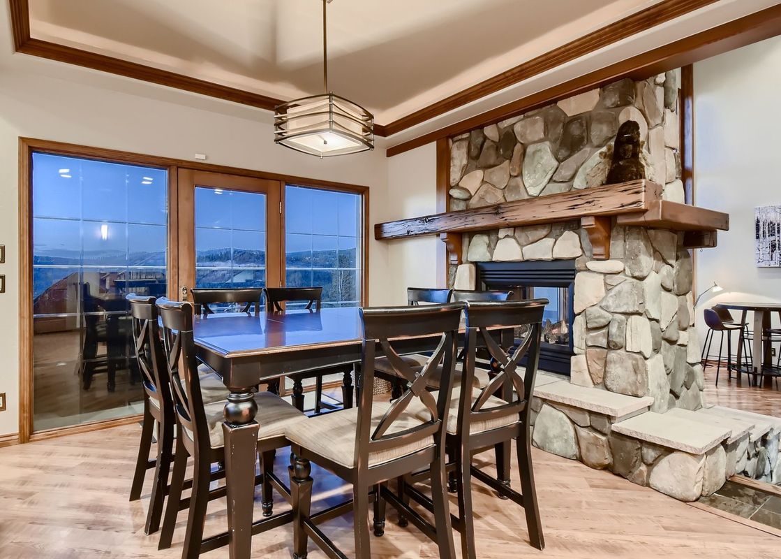 The dining area at Moose on the Loose features a large table with chairs, a stone fireplace, and windows showcasing a mountain view.