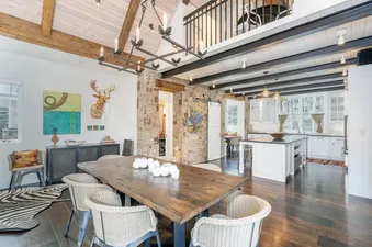 A large dining room with a long wooden table and woven chairs, leading into a modern white kitchen with an island and large windows.