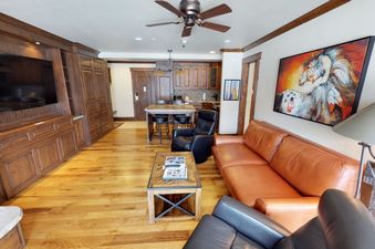 A modern living room with a leather sofa, a leather armchair, and a coffee table. The room has wooden floors, a large television on the wall, and a ceiling fan.