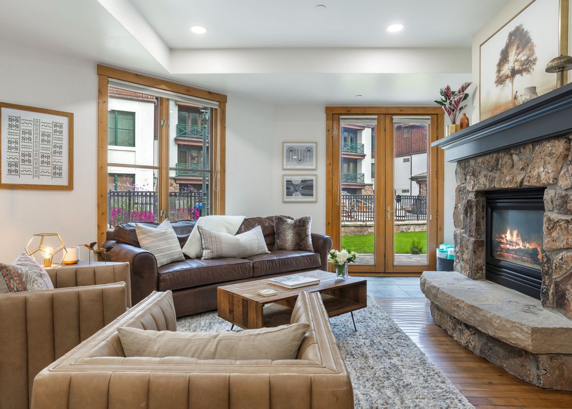 The living area at 118 Villa Montane features a plush leather sofa, two beige armchairs, a wooden coffee table, and a stone fireplace, all set against large windows with exterior views.