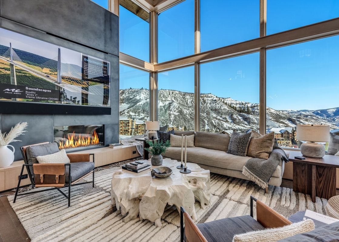 A luxurious Havens Haus living area with expansive mountain views, a large television above a modern fireplace, and comfortable seating around a unique coffee table.