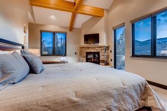 A spacious bedroom features a large bed with grey bedding, a stone fireplace with a television mounted above it, and multiple windows offering views of a snowy mountain landscape and pine trees.