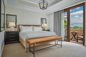 A spacious bedroom features a large bed with white linens and a wooden frame, complemented by black nightstands with lamps, a long leather bench at the foot of the bed, and a private balcony offering stunning mountain views.