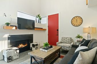 A bright and modern living room at Quarry A7 featuring a fireplace, a gray sofa, a wooden coffee table, a red door, and a large wooden clock.