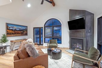 The living area of Le Chamonix Unit A features a large brown leather sofa, two green armchairs, a television mounted above a modern fireplace, and expansive windows offering a view of the snowy landscape.