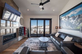 The living area of the Penthouse in the Pines features a cozy fireplace, a large mounted television, comfortable seating, and a stunning view of snow-covered mountains through large glass doors.