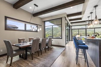 The dining room at Nine Peaks features a long wooden table with upholstered chairs, set against a backdrop of large windows showcasing mountain views. A stylish pendant light fixture hangs above the table, and a bar area with blue stools is visible to the