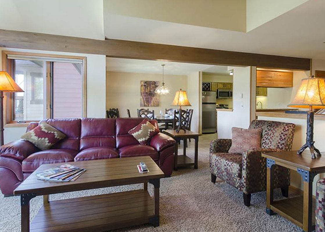 A cozy and spacious living room at the Ranch at Steamboat RA604, featuring a burgundy leather sofa, patterned armchairs, and rustic wooden furniture under warm lighting, with a dining area and kitchen visible in the background.