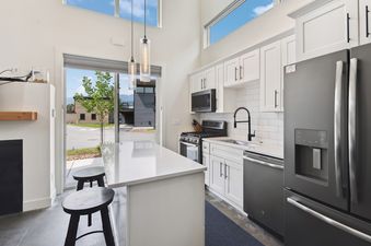 A modern kitchen in Quarry A4 features white cabinets, black appliances, a large island, and ample natural light from large windows offering views of the exterior.