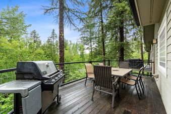 A spacious wooden deck at Suncrest Hideaway featuring a dining set and a BBQ grill, surrounded by a vibrant green forest under a clear blue sky.