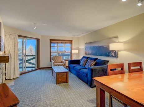 A cozy living area within the Blackcomb Springs One Bedroom Mountain View Suite features a blue sofa, a wooden coffee table, and a dining table, with large windows overlooking a snowy mountain landscape.