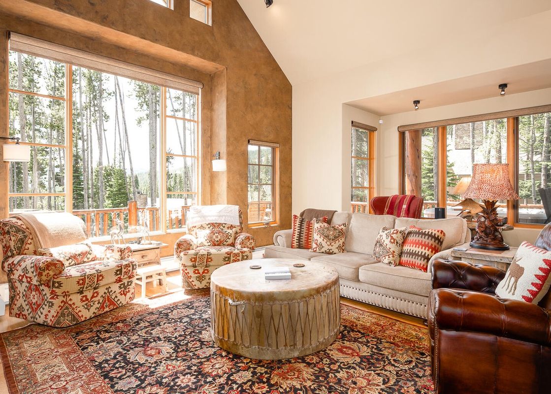 The living area at Moonlight Basin 4 Full Moon features a large sectional sofa with numerous throw pillows and a large rug, plus a unique drum coffee table. There is a wood-burning fireplace, and large windows provide stunning views of the mountains.