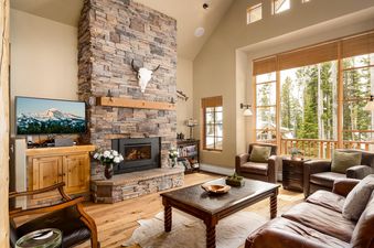 The living room of Moonlight Basin 5 Hidden Trail features a large stone fireplace, a wood-burning stove, leather furniture, and large windows with stunning views of the snowy mountain.