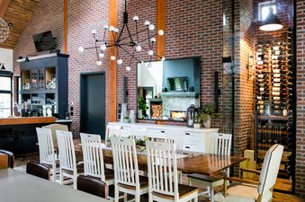 A large dining room with a long wooden table surrounded by chairs, a bar area, a fireplace with a TV above it, and a glass-enclosed wine rack.