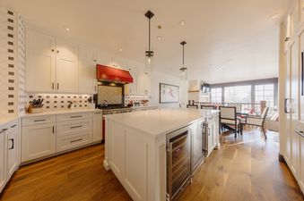 A spacious, well-lit kitchen featuring white cabinetry, a large kitchen island with built-in wine refrigerators, a red range hood above a gas stove, and warm wooden floors, opening into a dining area with large windows.
