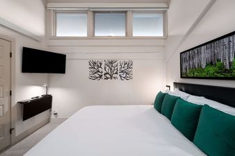 A bedroom with a large bed, white bedding, and green pillows. There is a black headboard, a flat-screen TV on the wall, and a large window.