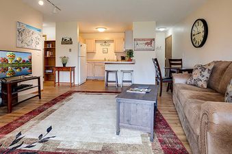 The Pines Condominiums P103C features an open living room with a brown sofa, a TV, a kitchen with a breakfast bar, and a dining area.