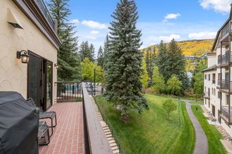 A spacious balcony at The Charter L022 with brick flooring, a BBQ grill, and chairs, offering scenic views of pine trees and mountains with vibrant autumn colors.
