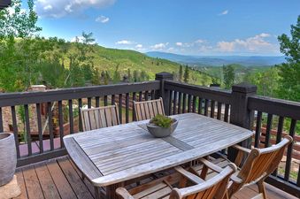 A wooden balcony with a dining table and chairs overlooks a vast green mountain landscape under a blue sky.