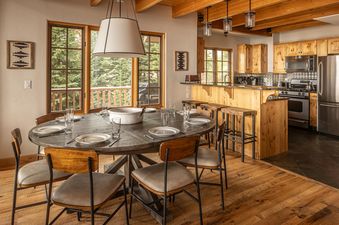 An open-concept living area at Moonlight Mountain 8 Happy Trails features a dining table and kitchen with views of evergreen trees outside.