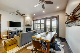 The living area and dining space are showcased in an open-concept layout at Blackstone Residence 1-B, featuring a stone fireplace, a large TV, a dining table, and a balcony view of the mountains.