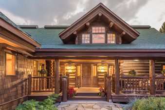 A rustic log cabin, the Huckleberry Hideaway, featuring a spacious front porch with warm lighting and comfortable seating.
