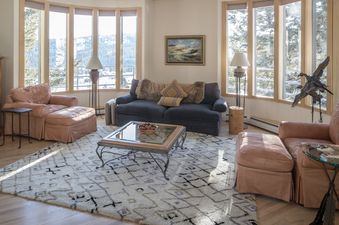 A cozy living room in Hillside Hideaway with plush seating and panoramic views of snow-covered mountains through large bay windows.