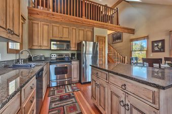 A fully equipped kitchen with wooden cabinetry, stainless steel appliances, granite countertops, and a patterned rug on a hardwood floor, with a loft visible above.