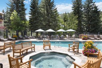 The image shows the outdoor pool and hot tub area at The Charter C1105, surrounded by wooden seating, lounge chairs, dining tables with umbrellas, and lush trees under a blue sky.
