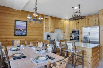 The dining and kitchen area at Comstock 203 features a long wooden dining table set for guests and an adjacent kitchen with light wood cabinetry and stainless steel appliances.