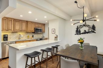 A bright, modern kitchen with stainless steel appliances and white cabinets, with a dining room and a view of the woods in the background
