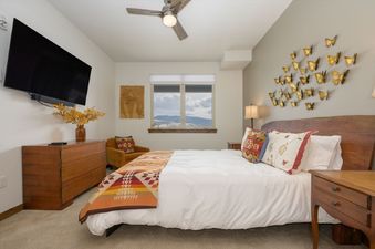 The bedroom at 448 Cranmer Unit 103 offers a large wall-mounted TV, a comfy bed with decorative pillows, a wooden dresser, and a window with mountain views.