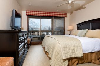 A cozy bedroom at The Gant Aspen 4 Bedroom Premier features a large bed, dark wood furniture, a television, and a sliding glass door leading to a balcony with mountain views.