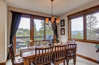 A dining room with a wooden table and chairs, a chandelier, and a view of trees from the windows.