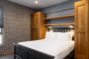 A comfortable king-sized bed with a dark wooden frame and white bedding, flanked by wooden built-ins and a textured stone wall.