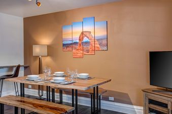 The dining area at Resort Plaza 5024 features a rustic wooden dining table with benches, set with white dishes and glasses, under an arched wall art.