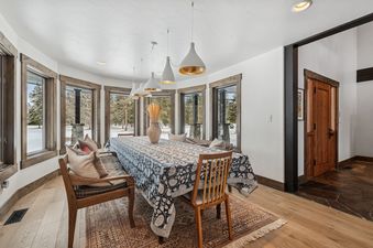 A cozy dining area features a wooden table with a patterned blue and white tablecloth, surrounded by large windows overlooking a snowy landscape with pine trees.
