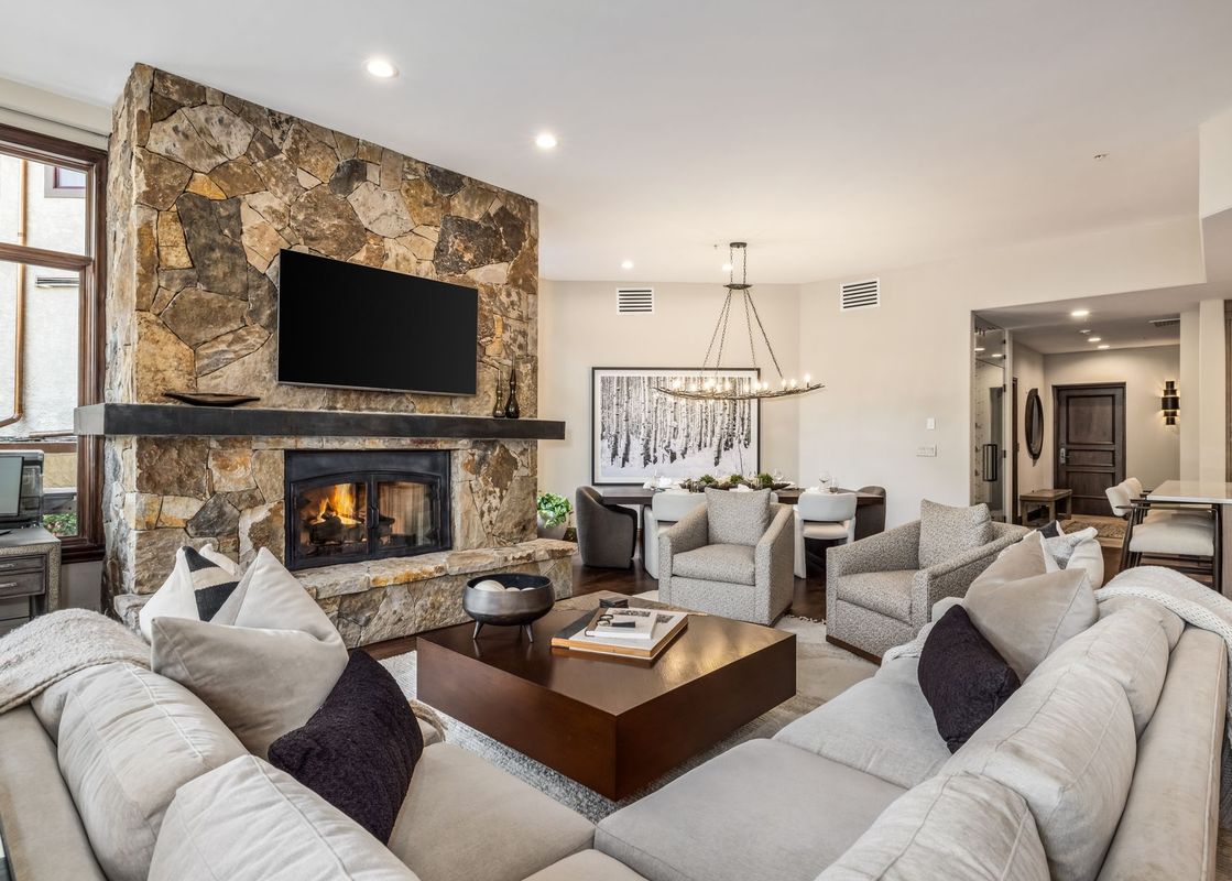 A cozy and elegant living room with a large stone fireplace, a sectional sofa, a coffee table, and an adjacent dining area.
