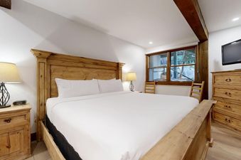 A cozy bedroom at Slope 101 with a wooden king-size bed, white linens, two nightstands with lamps, a large window, a dresser, and a wall-mounted TV.