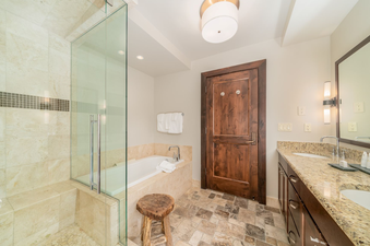 The bathroom in Lumiere Three Bedroom Luxury features a large glass shower, a jetted tub, a wooden stool, a double vanity, and a large mirror.