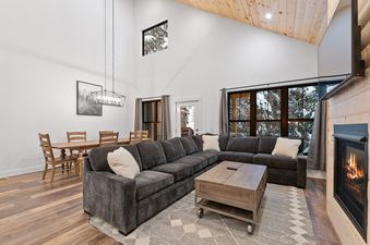 A large great room in Alpine Glow Ski Haus with a sectional sofa, coffee table, and a fireplace in the living area, and a wooden dining table with chairs under a modern chandelier in the dining area.