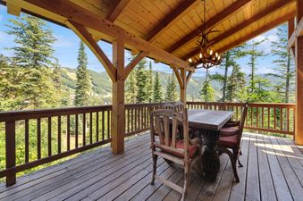 A rustic wooden deck with a dining table and chairs overlooks a mountain landscape with pine trees and ski slopes.