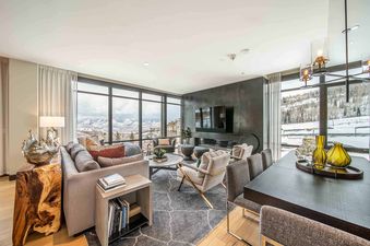 A luxurious and spacious lounge and dining area in One Snowmass Residence Club 407, featuring floor-to-ceiling windows with mountain views, a sectional sofa, a fireplace, a dining table, and modern decor.