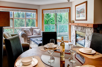 A cozy living area features a dining table set for guests, a comfortable armchair, and a fireplace, all bathed in natural light from large windows offering a view of lush greenery.
