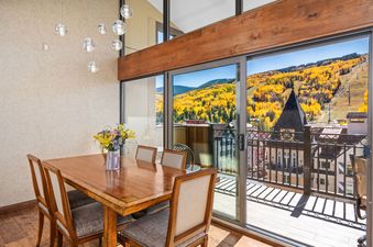 A dining room with a wooden table and chairs, illuminated by modern pendant lights, featuring large sliding glass doors that open to a balcony with a panoramic view of golden aspen trees on a mountain.