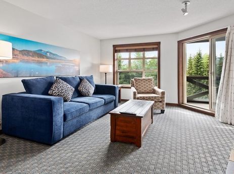 A cozy living room in Blackcomb Springs Two Bedroom Twins Suite with a blue sofa, a wooden coffee table, and a large window overlooking a forest.