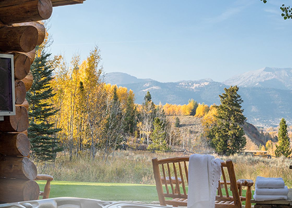 A private hot tub on a wooden deck offers stunning panoramic views of golden autumn trees and distant snow-capped mountains, complementing a rustic log cabin.