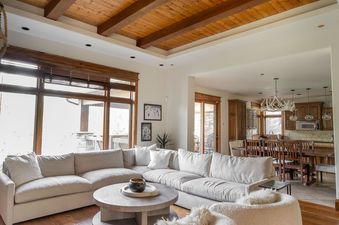 A spacious living room with a large, light-colored sectional sofa, a round wooden coffee table, and large windows with wooden blinds, showcasing a view into the adjacent kitchen and dining area.