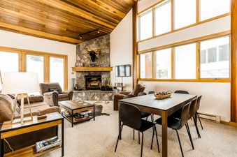 A cozy living room with a stone fireplace, comfortable seating, and a dining area, featuring large windows with an exterior view.