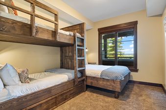 A cozy bedroom at Silver Star 1104 with a wooden bunk bed and an additional wooden bed, featuring a large window with a view of green trees and a bright blue sky.
